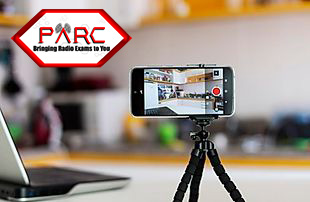 A phone held upright in a clip-style stand on a desk, positioned to view the candidate and their workspace during the exam.